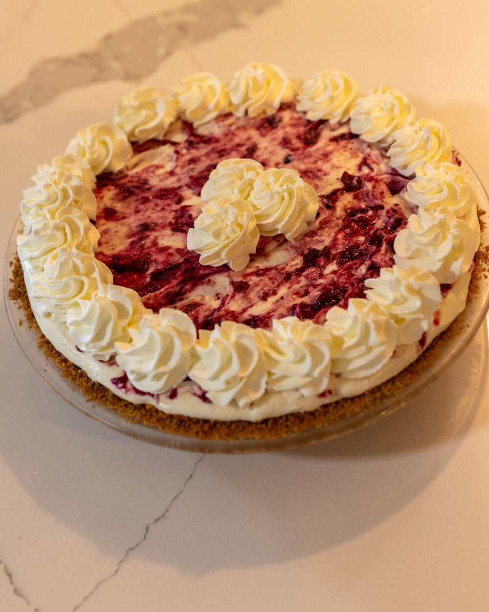 A mixed berry cream pie on a graham crust, ringed with piped whipped cream, cooling on a marble counter