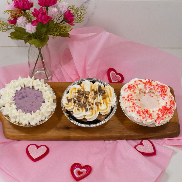 Three mini pies on a wooden board — berry, chocolate with caramel and candy pieces, and pink cream with sprinkles
